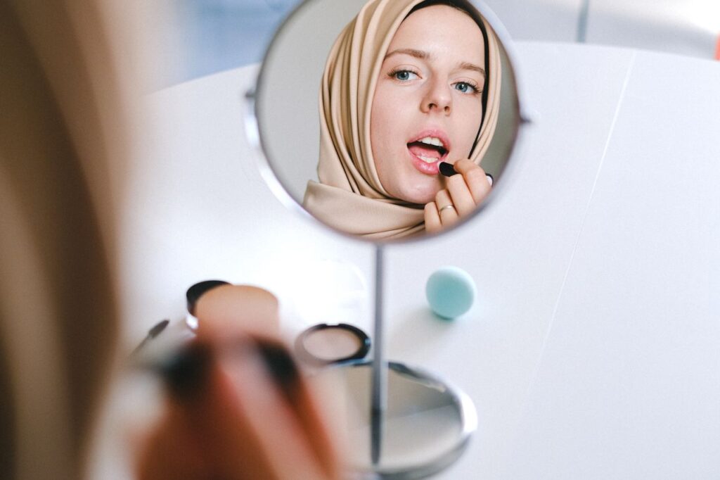 Muslim woman in hijab applies lipstick while looking at her reflection in a mirror.
